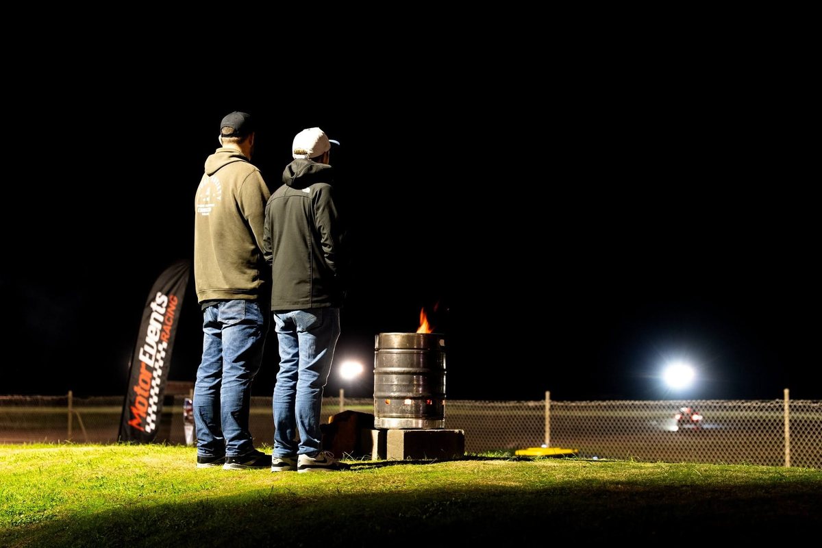 Two team members watching the race from the pit wall at night, warmed by a fire bucket.