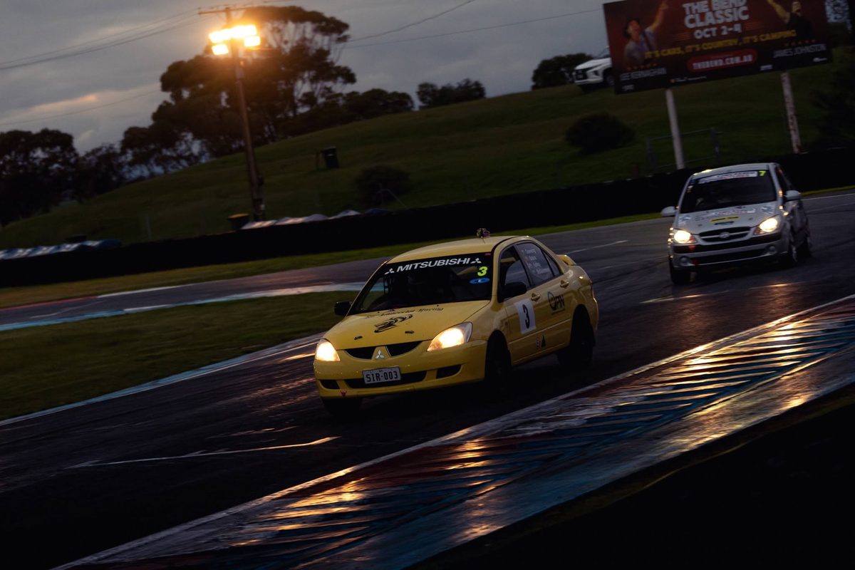 Two race cars on a wet circuit at dusk with headlights on, the leading car mid-corner.