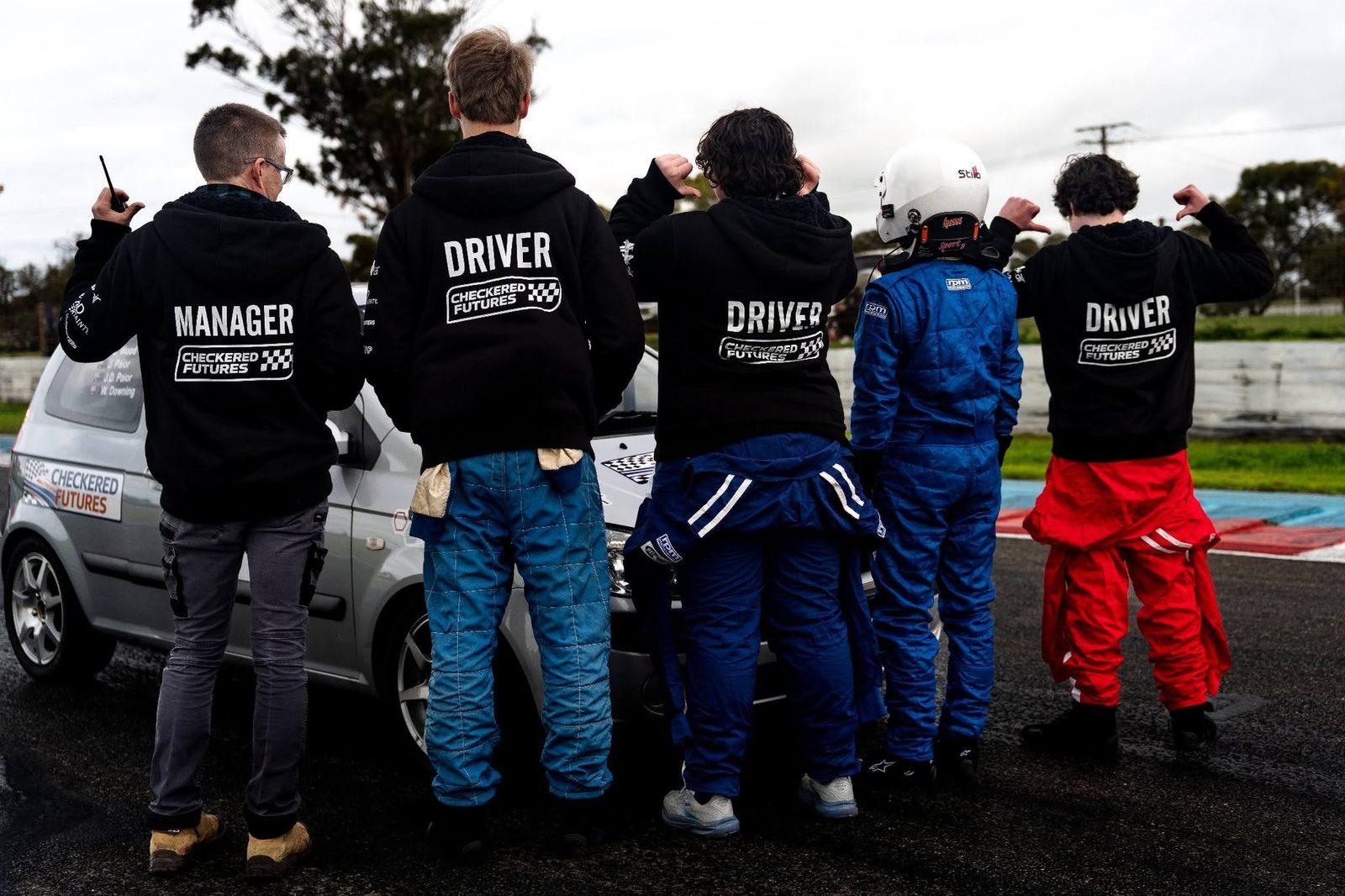 Five members of the Checkered Futures team stand with their backs to camera in Manager and Driver hoodies, facing their race car at the track.